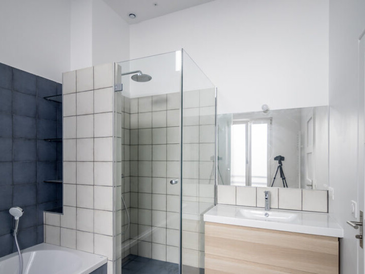 Salle de bain moderne avec cabine de douche en verre, lavabo avec vasque en bois et miroir au-dessus. Une baignoire est partiellement visible sur le côté gauche, mettant en valeur un mélange harmonieux de design contemporain et d'élégance haussmannienne suite à sa récente rénovation.