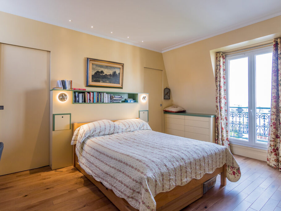 Une chambre bien éclairée évoque le charme de Montmartre avec un lit orné d'une courtepointe à rayures, une bibliothèque et des armoires intégrées derrière, un bureau et une chaise accueillants à gauche et une fenêtre avec des rideaux à motifs à droite, créant une atmosphère rappelant un Atelier d'artiste.