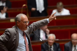 Un homme en costume fait des gestes tout en parlant à un podium dans un cadre formel, avec plusieurs personnes assises en arrière-plan.