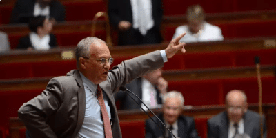 Un homme en costume fait des gestes tout en parlant à un podium dans un cadre formel, avec plusieurs personnes assises en arrière-plan.