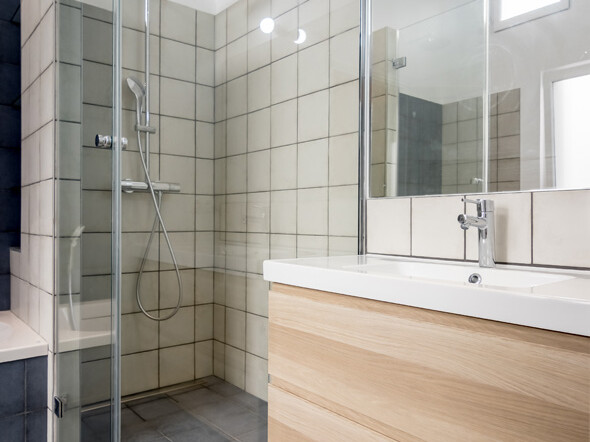 Salle de bain moderne avec douche vitrée, murs carrelés beiges, lavabo blanc avec meuble en bois et carrelage au sol à motifs incarnant un sens élégant de la rénovation dans le style haussmannien.