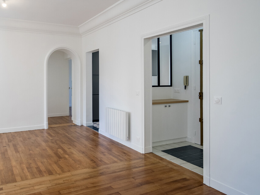 Un intérieur minimaliste avec parquet et murs blancs. Il comprend une porte en arc, un petit coin cuisine carrelé avec fenêtre et un couloir avec radiateur. Le charme subtil de cet espace reflète une rénovation élégante rappelant le style haussmannien classique.