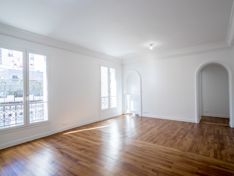 Chambre spacieuse et vide avec murs blancs, parquet en bois, grandes fenêtres et porte cintrée menant à une autre pièce. Cet espace haussmannien magnifiquement rénové allie charme historique et élégance moderne.