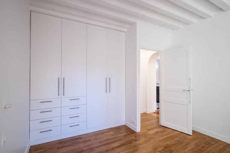 Un dressing blanc aux multiples tiroirs et armoires est installé dans une pièce au parquet, soulignant une rénovation récente. Une porte ouverte dévoile une pièce adjacente, ajoutant au charme du style haussmannien.