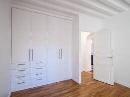 Un dressing blanc aux multiples tiroirs et armoires est installé dans une pièce au parquet, soulignant une rénovation récente. Une porte ouverte dévoile une pièce adjacente, ajoutant au charme du style haussmannien.