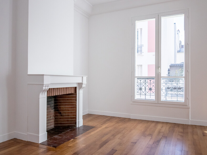 Une pièce lumineuse et vide avec un parquet en bois, une cheminée blanche sur le mur de gauche et une grande fenêtre sur la droite avec vue sur les bâtiments voisins. L'espace respire le charme haussmannien classique, attendant votre touche personnelle pour une rénovation parfaite.