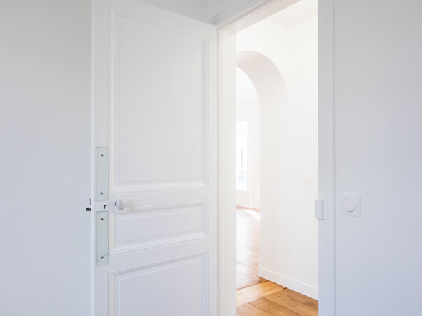 Une porte blanche s'ouvre sur une pièce lumineuse aux murs blancs et au parquet en bois, capturant le charme d'une rénovation haussmannienne.