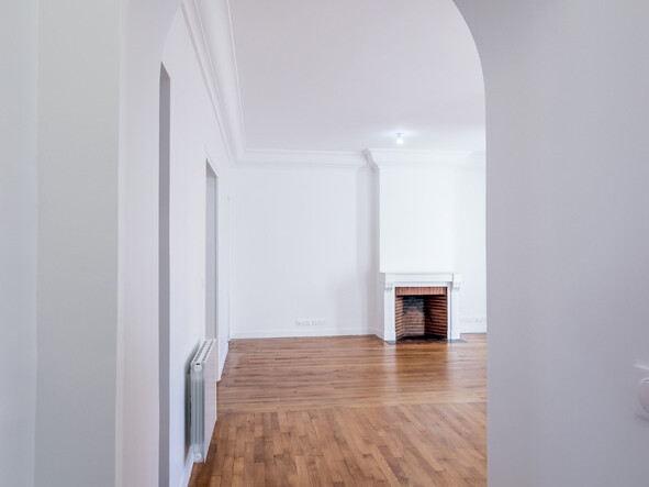 Un couloir au sol en bois mène à une pièce blanche agrémentée d'une cheminée, mettant en valeur un élégant style haussmannien. Les murs et le plafond, également blancs, témoignent du soin apporté à la rénovation.