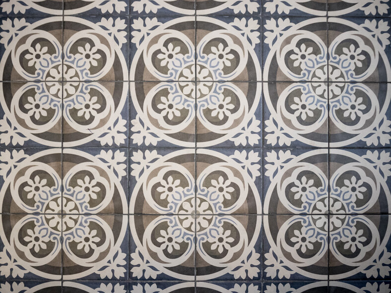 Un sol avec un motif répétitif de motifs floraux et circulaires blancs complexes sur des carreaux bleus et gris, rappelant l'élégance haussmannienne intemporelle, parfait pour votre prochain projet de rénovation.