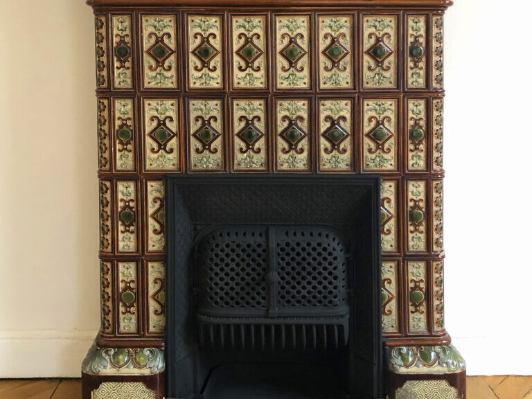 Une cheminée vintage ornée de motifs de carreaux complexes et d'une protection avant noire, placée contre un plancher en bois et un mur blanc cassé, capturant parfaitement l'élégance du charme haussmannien.