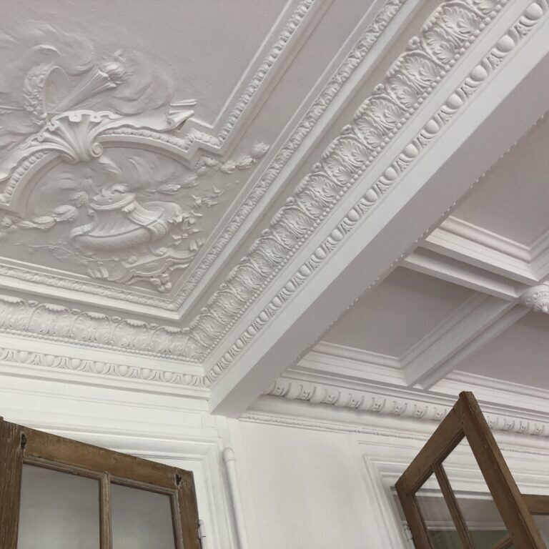 Un plafond blanc orné de détails de plâtrerie complexes et de moulures couronnées, rappelant l'architecture haussmannienne, accompagné de fenêtres à cadre en bois partiellement ouvertes.