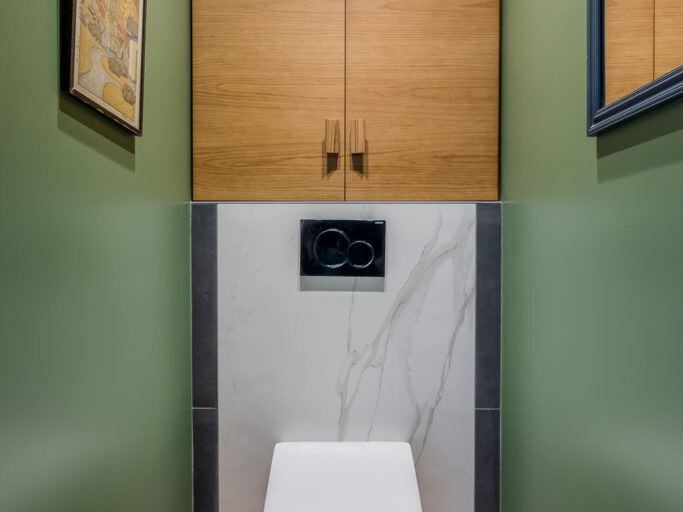 Une petite salle de bain moderne aux murs verts rappelant les charmantes rues de Montmartre, des armoires en bois et des toilettes suspendues. Deux tableaux encadrés font écho au style d'un atelier d'artiste, ajoutant une touche d'élégance artistique à l'espace.