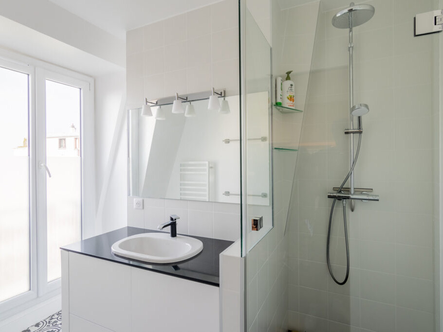 Une salle de bain moderne avec un lavabo élégant, un grand miroir, une cabine de douche en verre et un pommeau de douche mural. La pièce est lumineuse grâce à la lumière naturelle provenant d'une grande fenêtre, rappelant un atelier d'artiste à Montmartre.