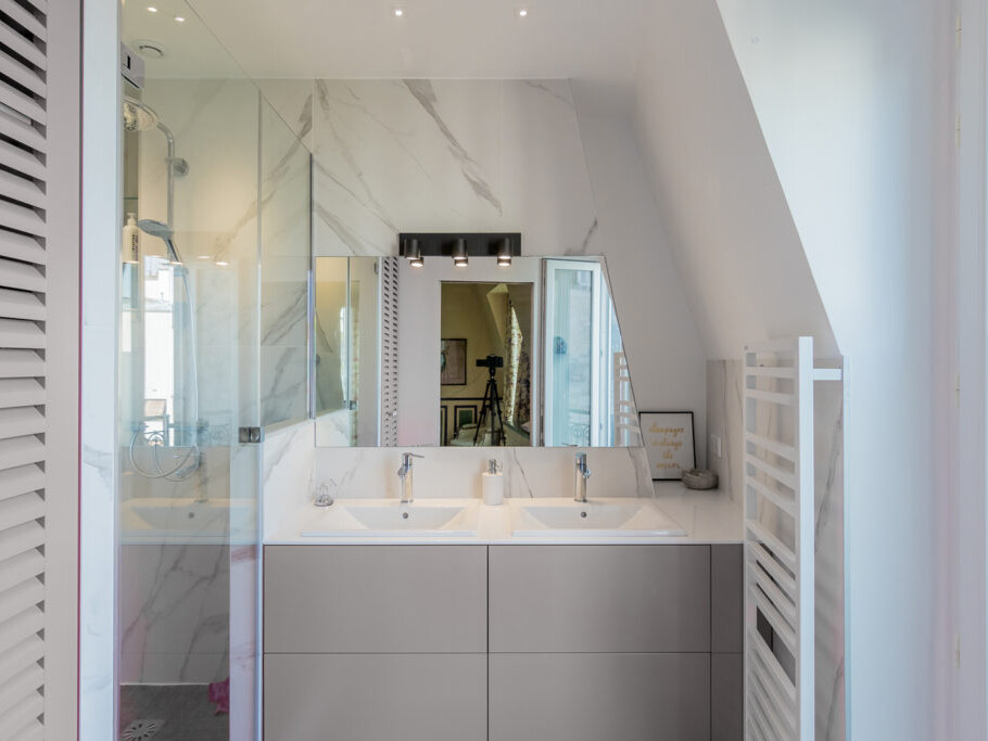 Une salle de bain moderne inspirée du charme artistique de Montmartre, avec un meuble-lavabo double, un grand miroir, une douche vitrée et une fenêtre. Les murs sont blancs avec des accents de marbre subtils, rappelant la simplicité élégante d'un atelier d'artiste.