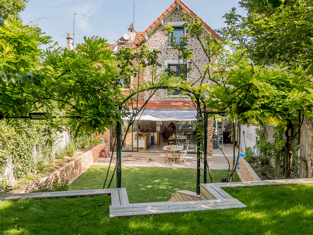 Une maison à deux étages avec un toit rouge et une façade en pierre se dresse au bout d'une pelouse bien entretenue, partiellement couverte par une pergola avec des plantes grimpantes. La maison dispose d'un patio ouvert avec des sièges extérieurs et d'une extension récente pour un espace de vie supplémentaire.