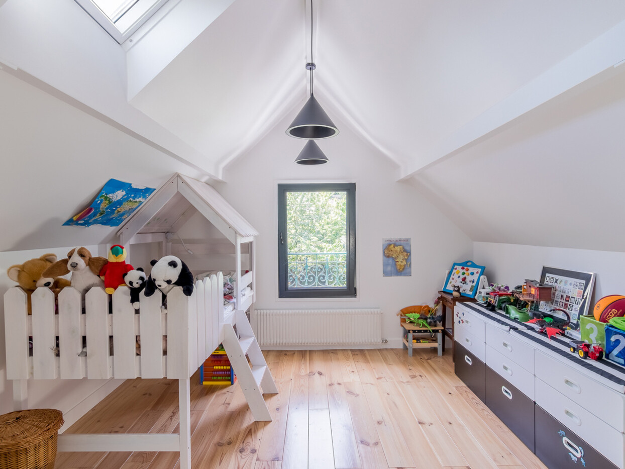 Chambre d'enfant dans un grenier surélevé avec un plancher en bois. La pièce comprend un petit lit avec un cadre de maisonnette, des animaux en peluche, des jouets et des meubles de rangement blancs le long du mur. Une lucarne et une fenêtre apportent de la lumière naturelle.