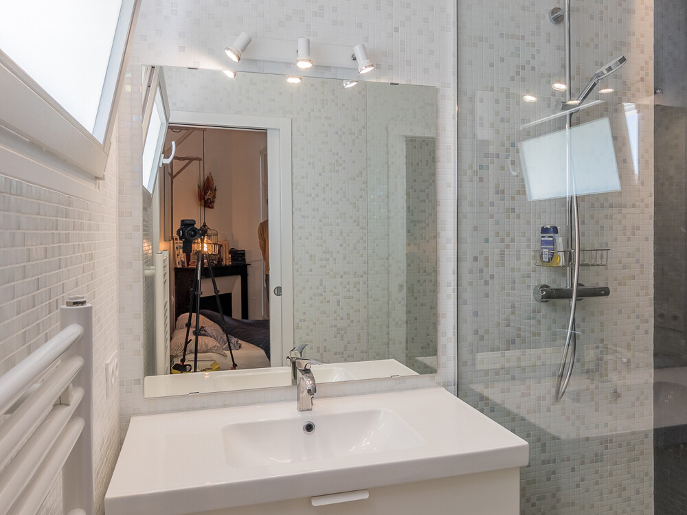 Salle de bains minimaliste avec meuble-lavabo blanc, miroir mural et douche vitrée avec pommeau de douche. Un radiateur sèche-serviettes ajoute de la fonctionnalité. Dans le reflet du miroir, indiquant un projet d'extension, on peut voir une caméra sur un trépied.