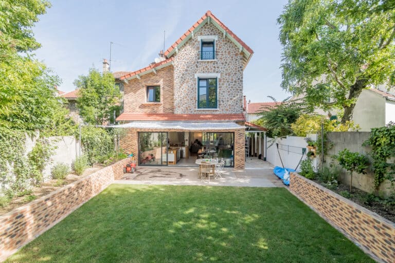 Une maison en brique de trois étages avec une porte-fenêtre vitrée, un coin salon extérieur et une pelouse bien entretenue est entourée d'arbres et de verdure. La surélévation récente ajoute un charme supplémentaire à cette demeure chaleureuse.