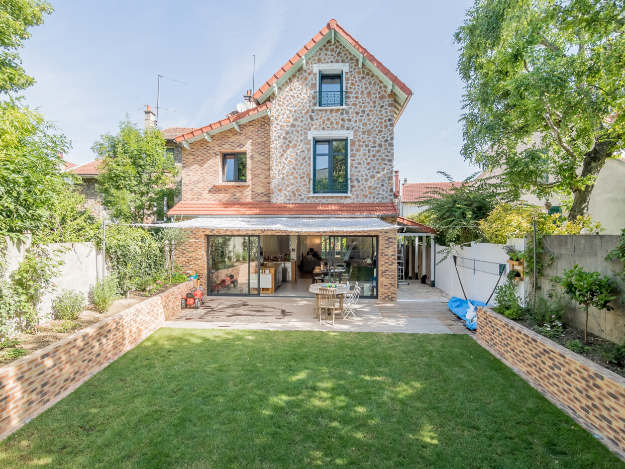 Une maison en brique de trois étages avec une porte-fenêtre vitrée, un coin salon extérieur et une pelouse bien entretenue est entourée d'arbres et de verdure. La surélévation récente ajoute un charme supplémentaire à cette demeure chaleureuse.