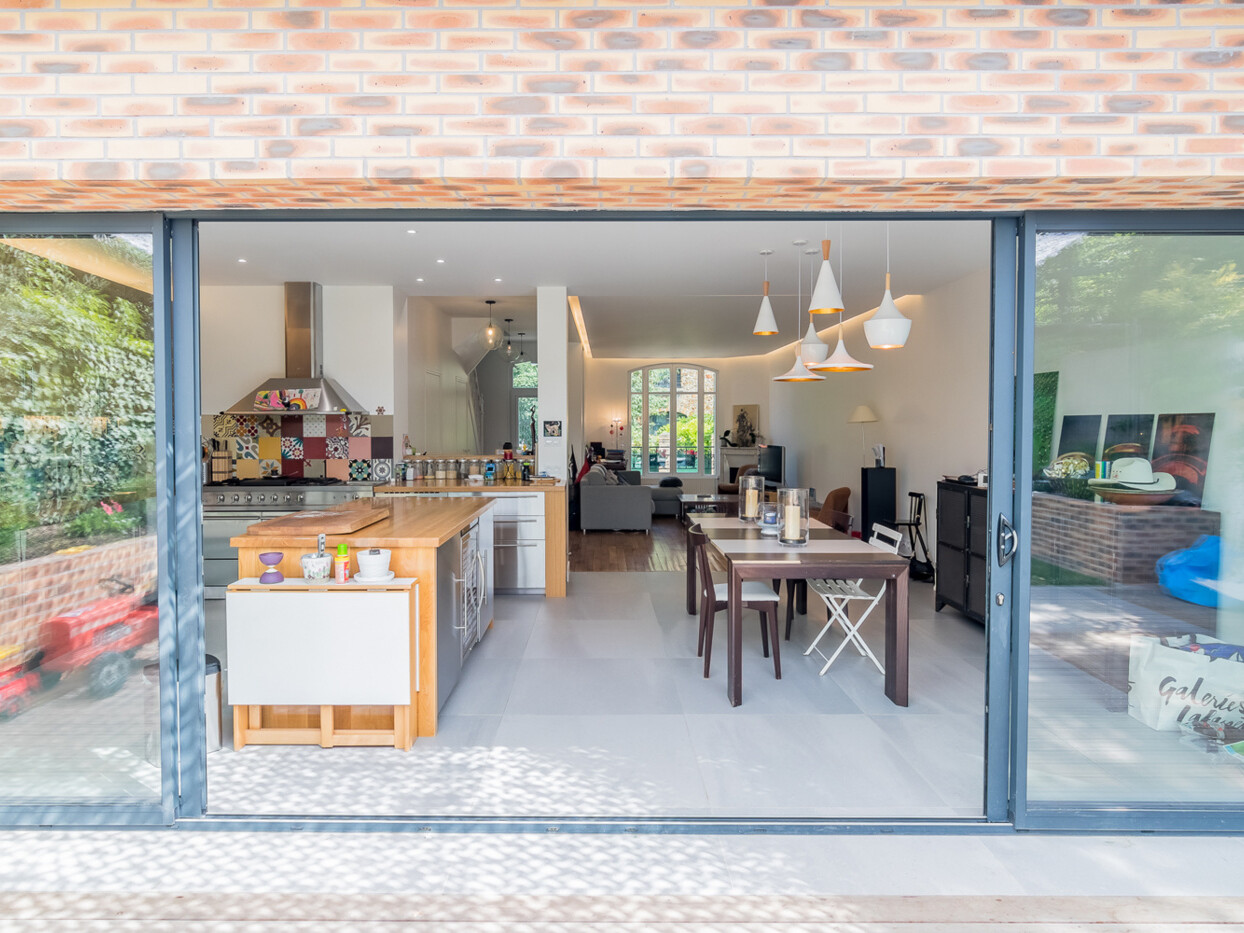 Vue d'une cuisine et salle à manger modernes et ouvertes avec un extérieur en brique. De grandes portes coulissantes en verre s'ouvrent pour révéler l'intérieur bien éclairé et spacieux, faisant partie d'une extension élégante.