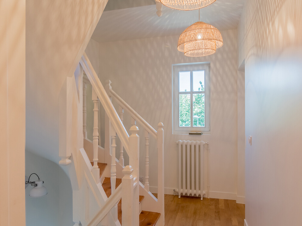 Un couloir lumineux avec parquet et murs blancs, avec un escalier et deux luminaires suspendus en osier, met en valeur une élégante surélévation. Une fenêtre permet à la lumière naturelle de pénétrer dans la zone agrandie.