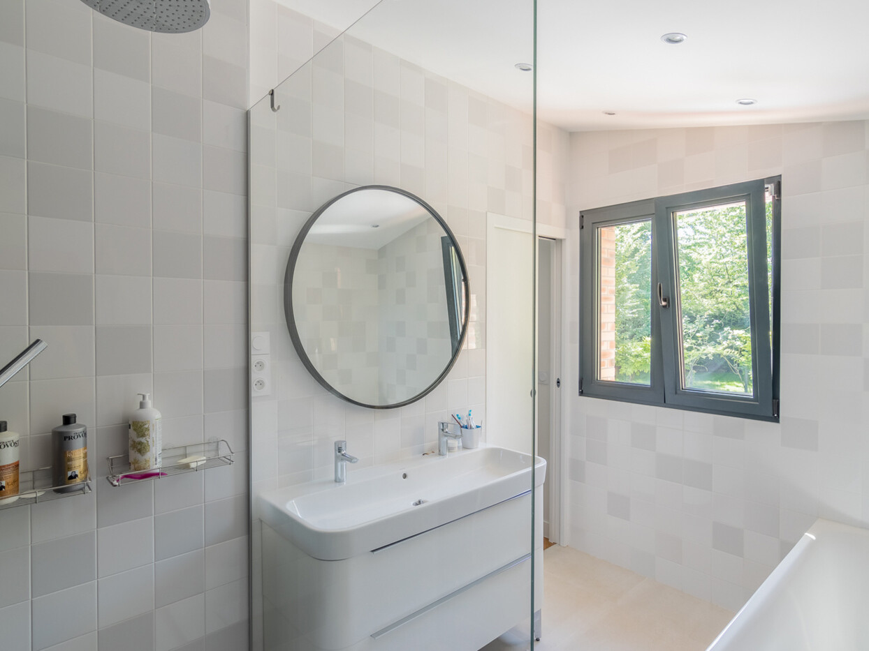 Salle de bain moderne avec un miroir circulaire au dessus d'un lavabo blanc, un espace douche avec produits de toilette à gauche et une fenêtre à droite laissant entrer la lumière naturelle provenant de l'extension récente.