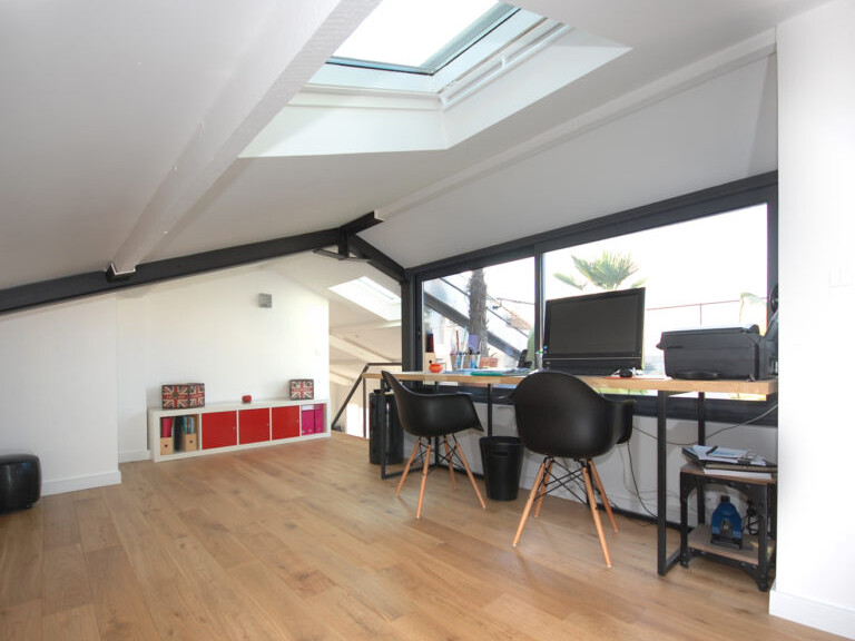 Un bureau mansardé moderne avec une lucarne, un parquet en bois, un grand bureau avec un ordinateur, des livres et deux chaises noires. Une étagère avec des boîtes de rangement est contre le mur incliné à gauche. Parfaitement aménagé au sein des combles, cet espace cosy offre à la fois fonctionnalité et style.
