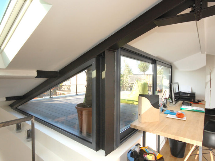 Un bureau mansardé moderne, astucieusement aménagé dans les combles avec un toit en pente, dispose d'une grande fenêtre, d'un bureau en bois, d'une chaise et de fournitures de bureau. La fenêtre offre une vue sur une terrasse extérieure ornée de plantes en pot et de meubles.