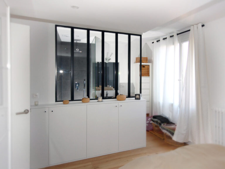 Une chambre moderne avec une grande fenêtre, des rideaux blancs, un lit et une armoire blanche encastrée. Une cloison avec des panneaux de verre sépare la pièce d'un espace adjacent. Le parquet en bois clair ajoute de la chaleur à l'espace, intégrant harmonieusement l'esthétique d'un aménagement de combles aménager.