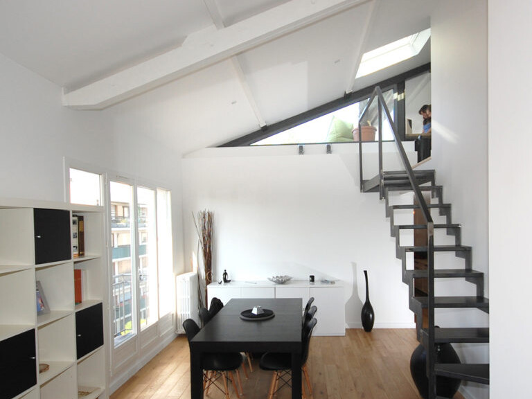 Un loft moderne avec un plancher en bois, une table à manger noire, une étagère ouverte et un escalier en métal menant à un niveau supérieur, offre un espace idéal pour aménager. La conception garantit que chaque coin est utilisé efficacement et pourrait être parfait pour intégrer une terrasse élégante.