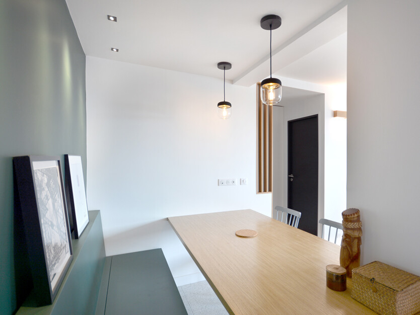 Salle à manger minimaliste avec une table en bois, trois chaises blanches, deux luminaires suspendus, des œuvres d'art encadrées sur un mur d'accent vert et des objets décoratifs sur la table. Les rénovations mettent en valeur le mélange parfait de simplicité et d'élégance dans cet espace deux pièces rafraîchi.