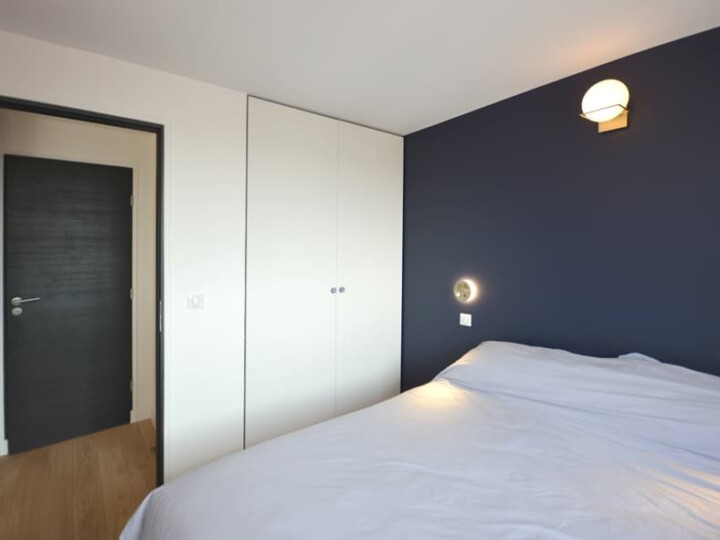 Une chambre moderne avec un mur d'accent bleu, un grand lit avec des draps blancs, une armoire intégrée et un plancher en bois qui met en valeur la rénovation élégante de la pièce. La porte mène au reste du charmant deux pièces.