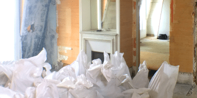 Une pièce en cours de rénovation avec des murs sablés, une cheminée couverte et des draps blancs de protection drapés sur les objets. Une porte mène à une autre pièce peu meublée à l'arrière-plan.