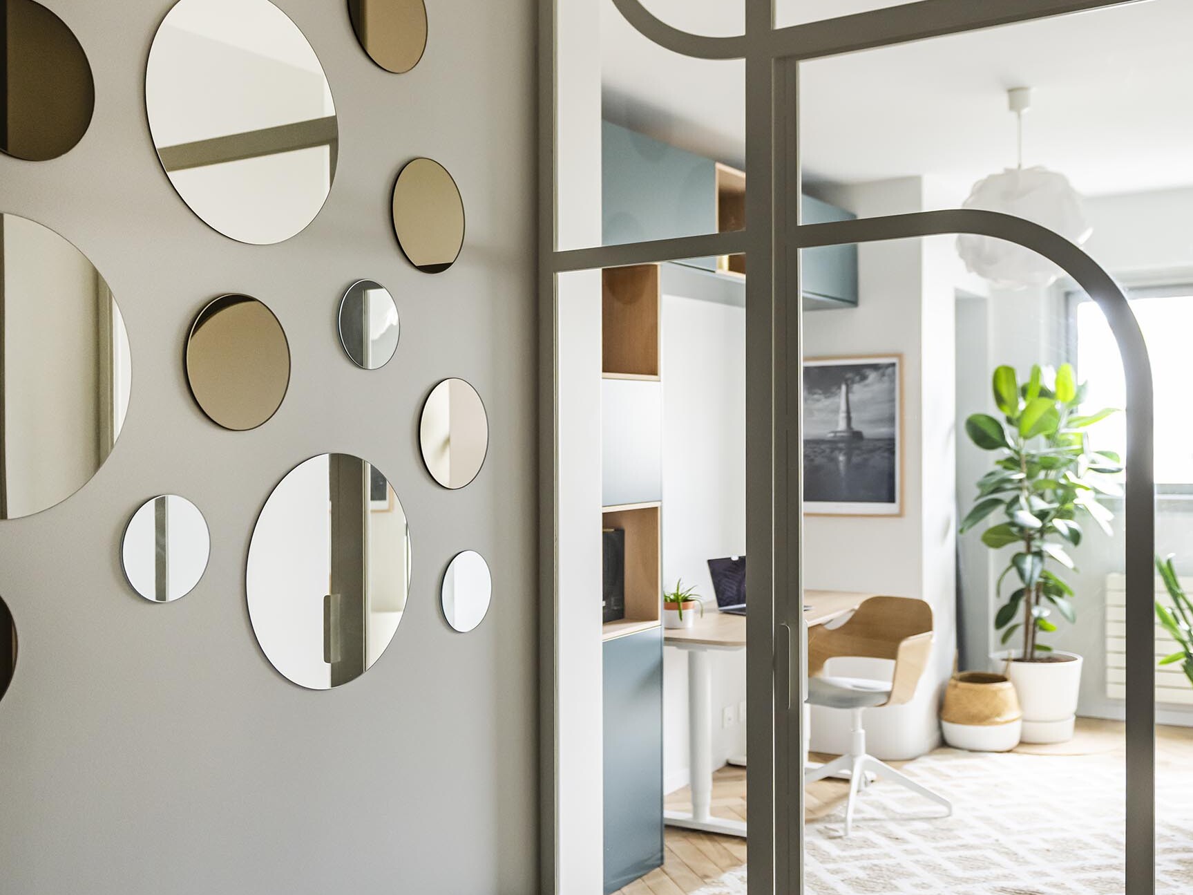 Intérieur moderne montrant une pièce élégante avec un mur de miroirs ronds, un banc à motifs géométriques, un espace de travail avec des étagères et un coin salon avec des plantes près d'une grande fenêtre.