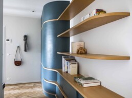 Un couloir moderne est doté d'un meuble de rangement bleu incurvé avec des étagères en bois enveloppantes contenant des livres et des bibelots. Un plancher en bois franc à chevrons s'étend à travers l'espace et un porte-manteau peut contenir un sac et une écharpe.