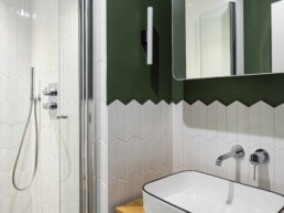 Une salle de bain moderne avec des carreaux hexagonaux blancs, une partie murale verte, un lavabo rectangulaire avec robinet mural et un espace douche vitré.