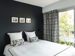 Une chambre moderne comprend un lit soigneusement fait avec des draps blancs et des coussins à motifs géométriques, un mur d'accent sombre avec des images encadrées et une porte coulissante en verre menant à un balcon avec des sièges extérieurs.