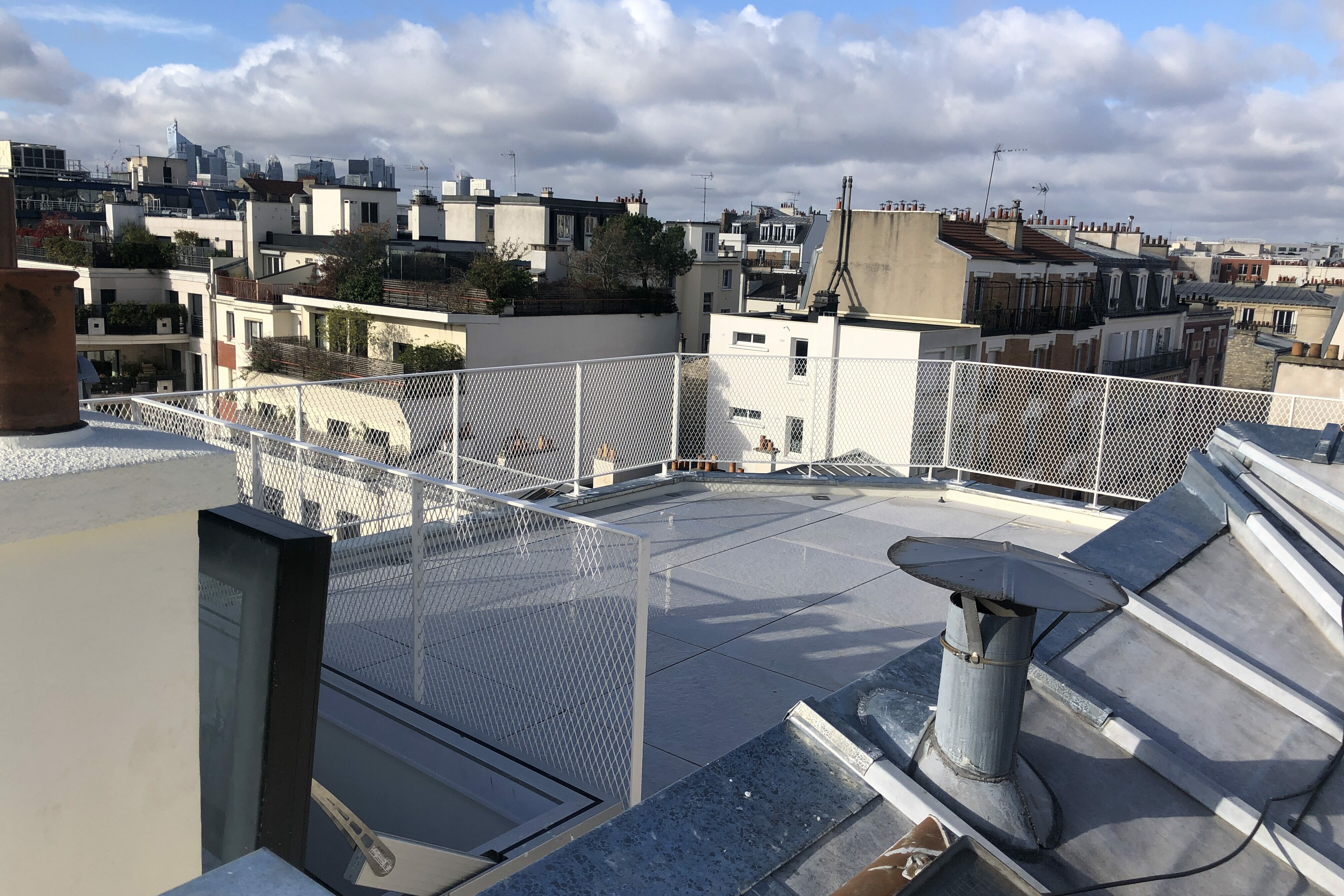 Vue sur les toits d'un paysage urbain avec un mélange de bâtiments modernes et anciens, un ciel nuageux et une clôture métallique au premier plan.