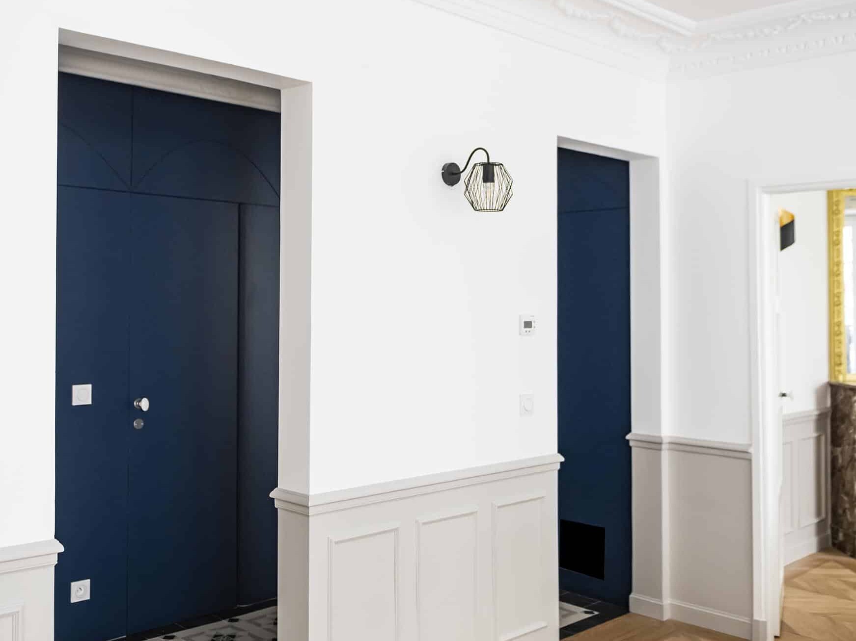 Le couloir, qui rappelle le style haussmannien, est agrémenté de deux portes bleues aux moulures décoratives. Le sol à chevrons renforce son élégance, tandis qu'une applique murale illumine doucement l'espace entre les portes, suggérant peut-être une chambre en plus au-delà.