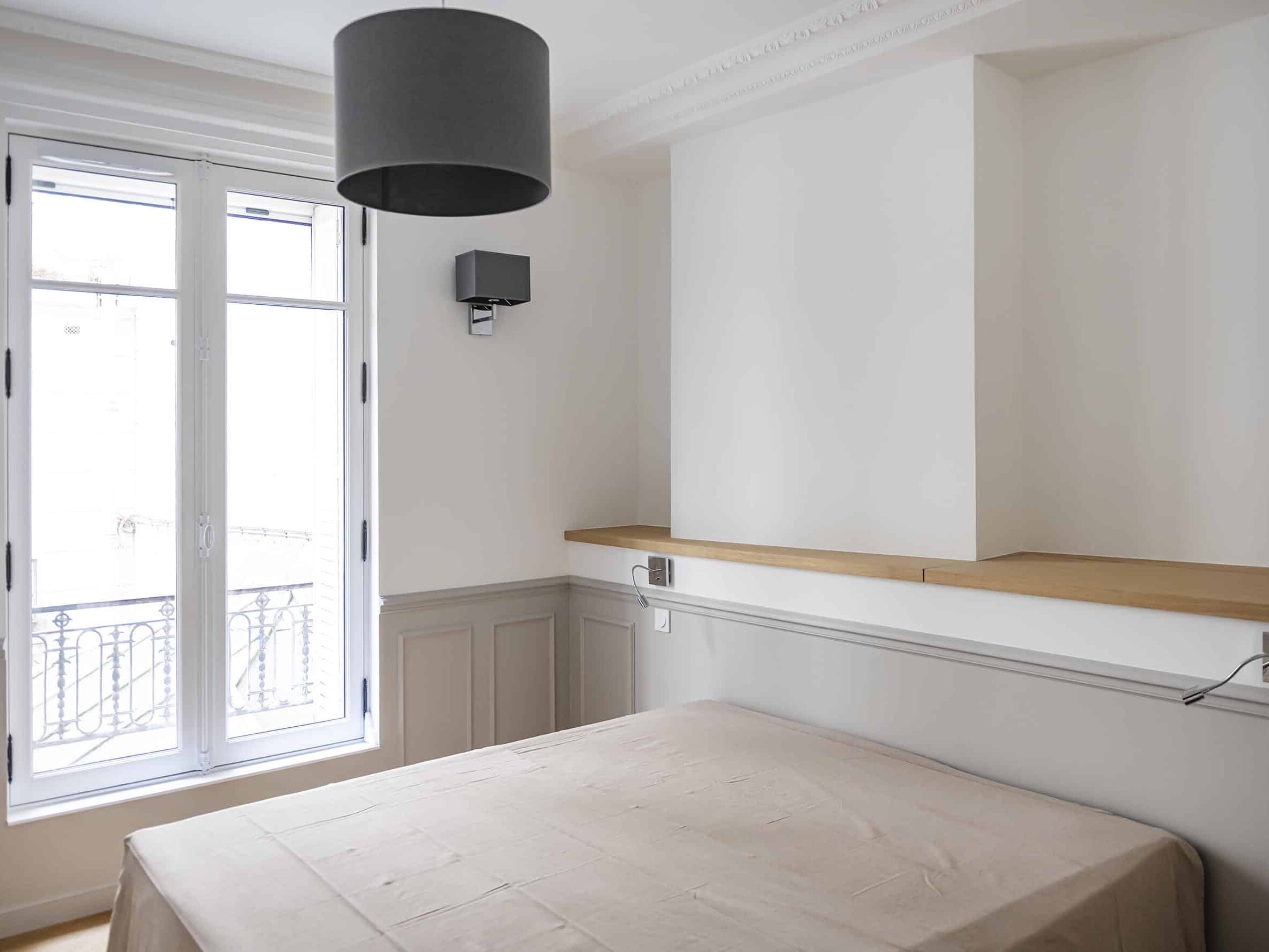 Une chambre minimaliste avec un grand lit, des murs gris clair et une fenêtre offrant une vue exquise. Un plafonnier noir est suspendu au-dessus et une étagère en bois orne un mur. Cet espace serein ressemble à une chambre en plus nichée dans le charme de son environnement.