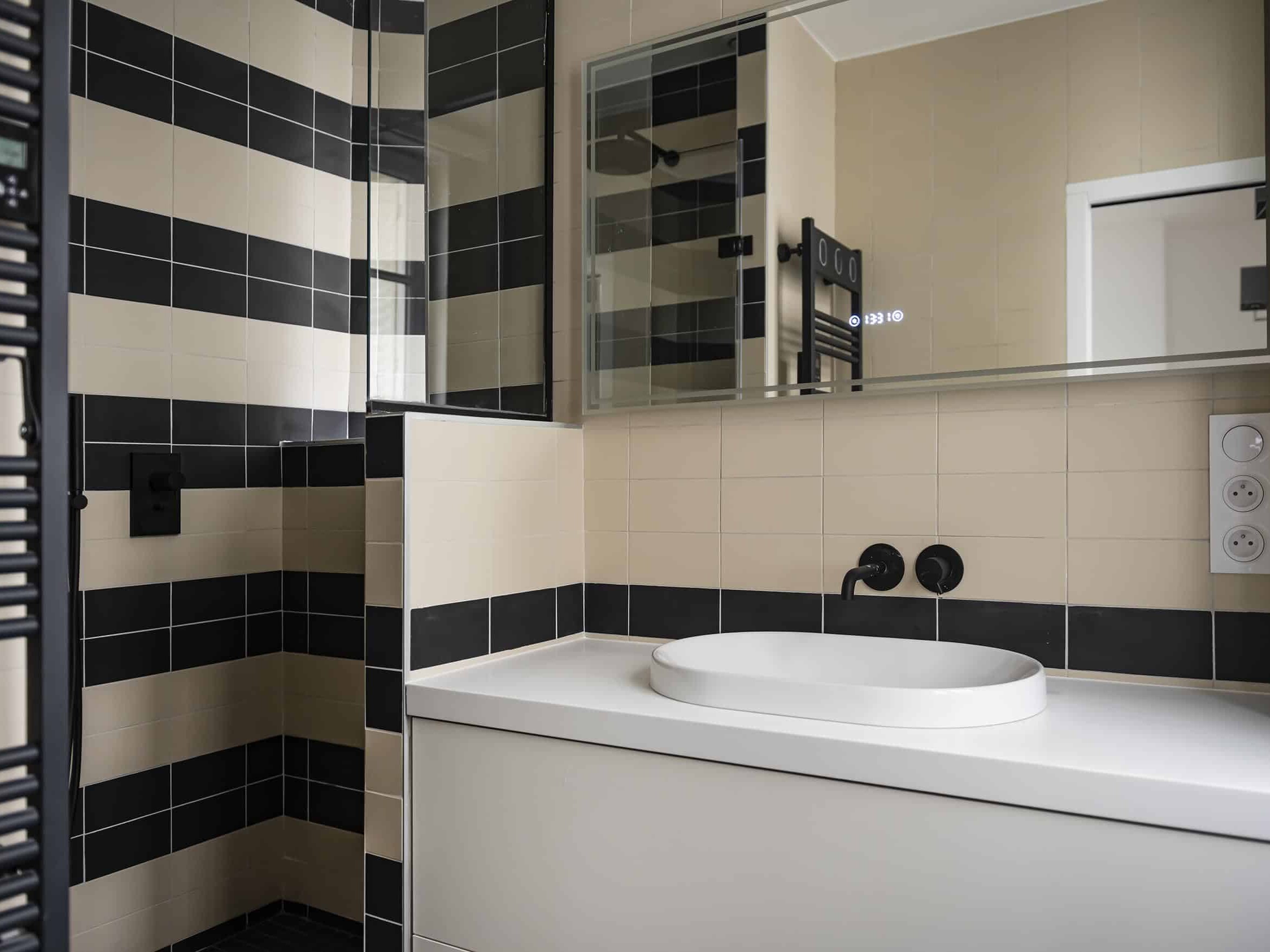 Salle de bain moderne avec un lavabo blanc sur meuble, entouré de carrelage noir et beige, rappelant un style haussmannien. Un miroir est au-dessus du lavabo et un radiateur sèche-serviettes noir est visible sur le mur de gauche, offrant une chambre en plus pour rehausser votre espace de vie.