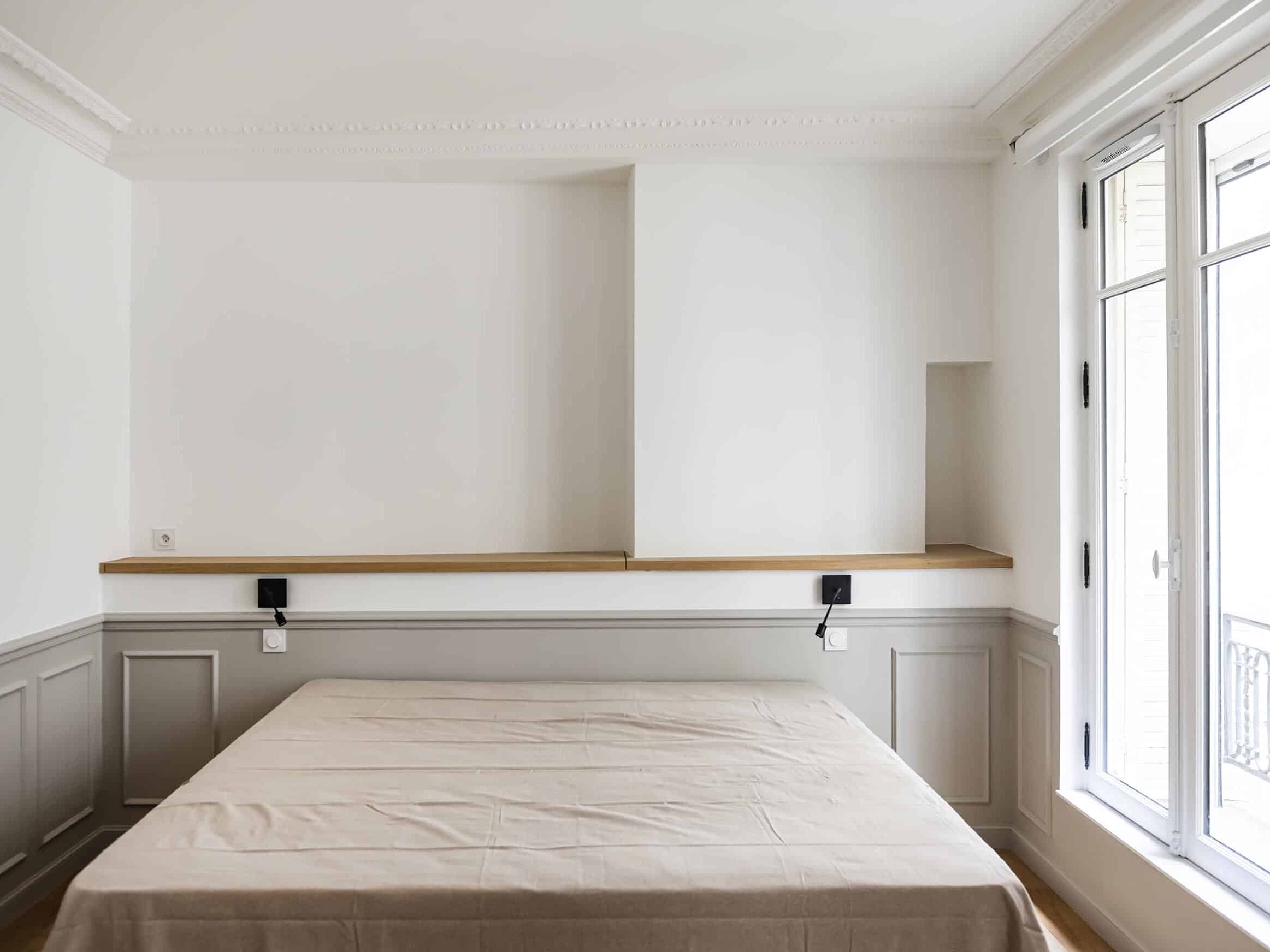 Une chambre minimaliste avec un lit beige, des murs blancs et de grandes fenêtres offrant une vue sur le balcon évoque l'élégance du style haussmannien. Des boiseries et des appliques murales ornent la tête de lit, créant une touche supplémentaire de charme classique.