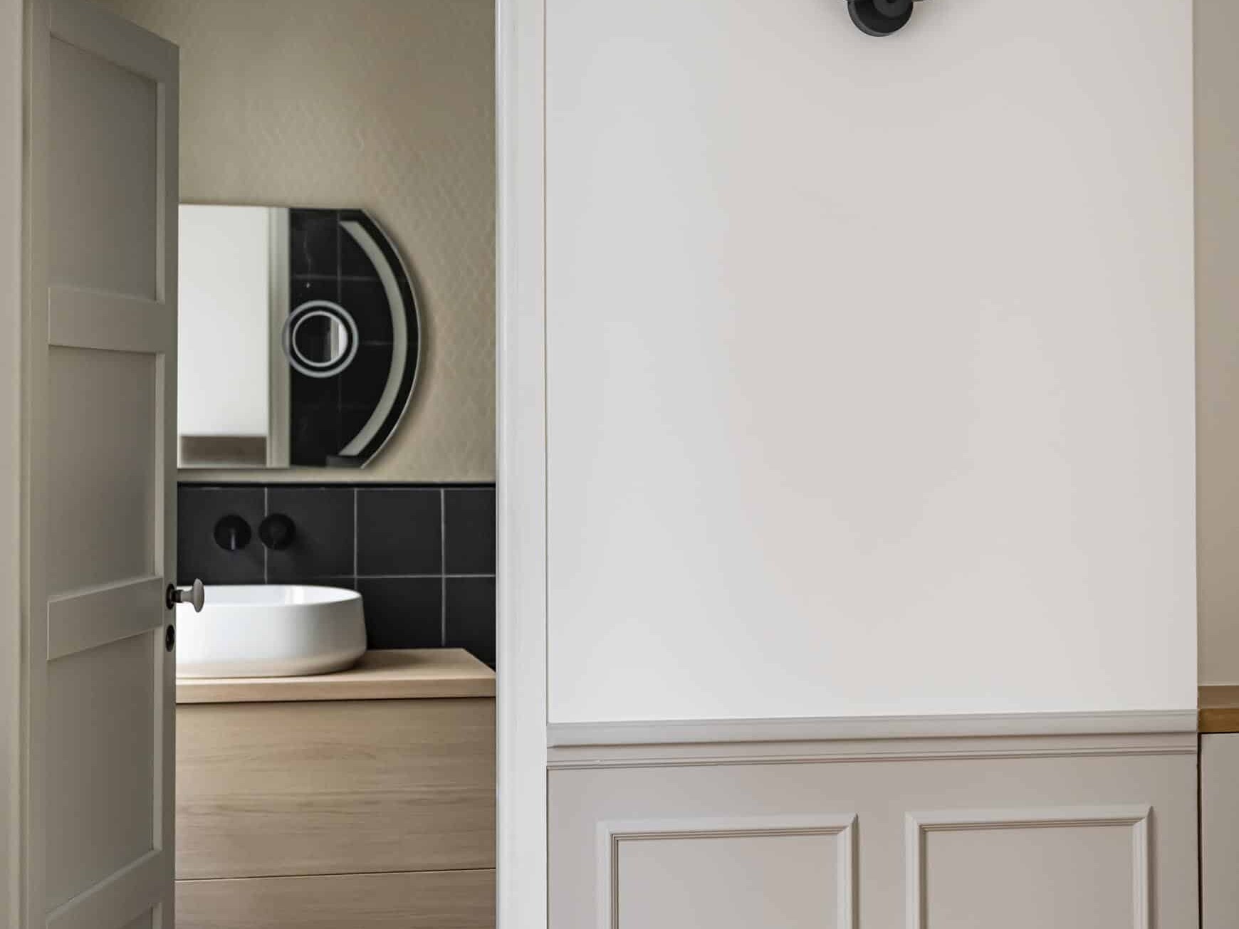 Une salle de bain vue à travers une porte ouverte présente un lavabo avec un miroir rond, un sol en carrelage à motifs et un luminaire moderne sur le mur adjacent, faisant écho à l'élégance d'une chambre en plus de style haussmannien.