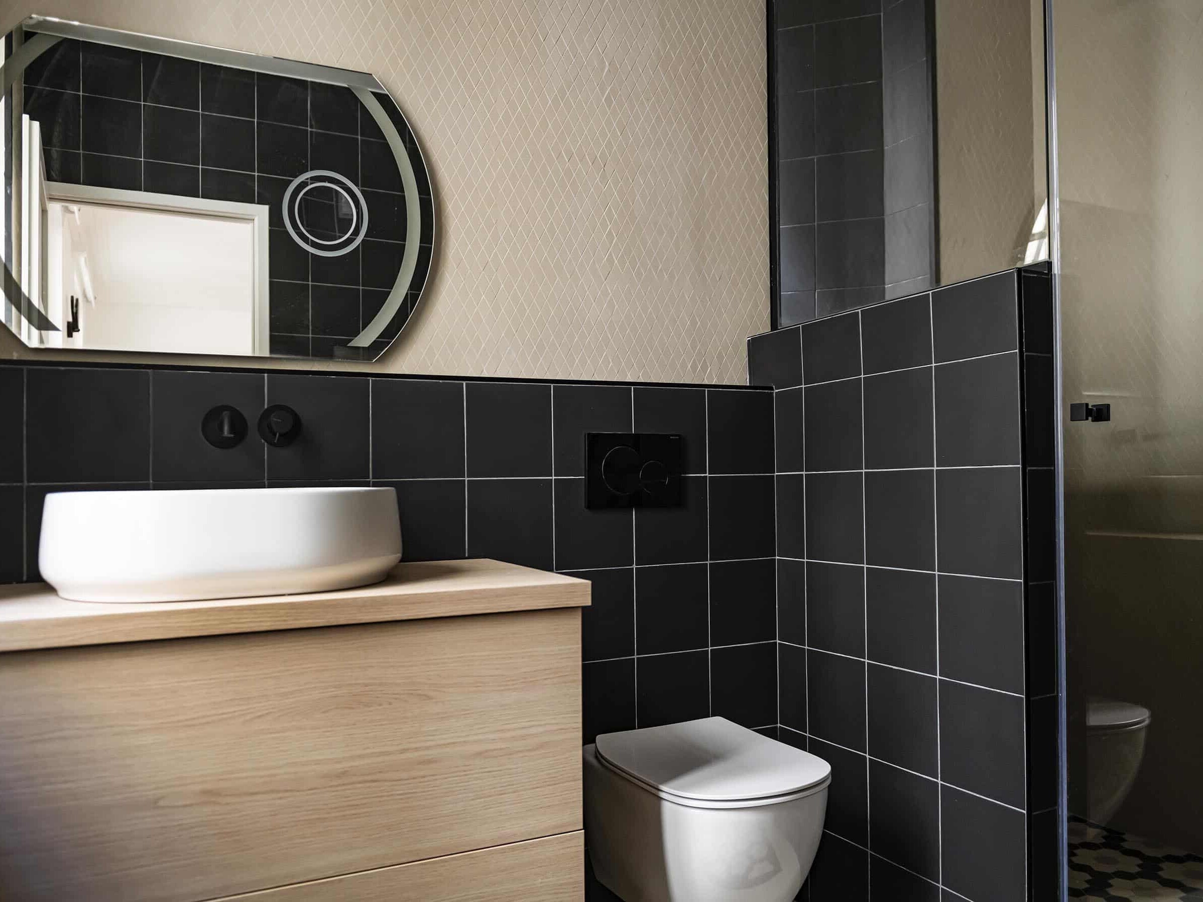 Une salle de bain moderne avec des murs carrelés noirs, un miroir rond et un lavabo blanc sur une vanité en bois complète parfaitement l'élégance haussmannienne de la maison, offrant une fonctionnalité proche de l'ajout d'une chambre en plus.