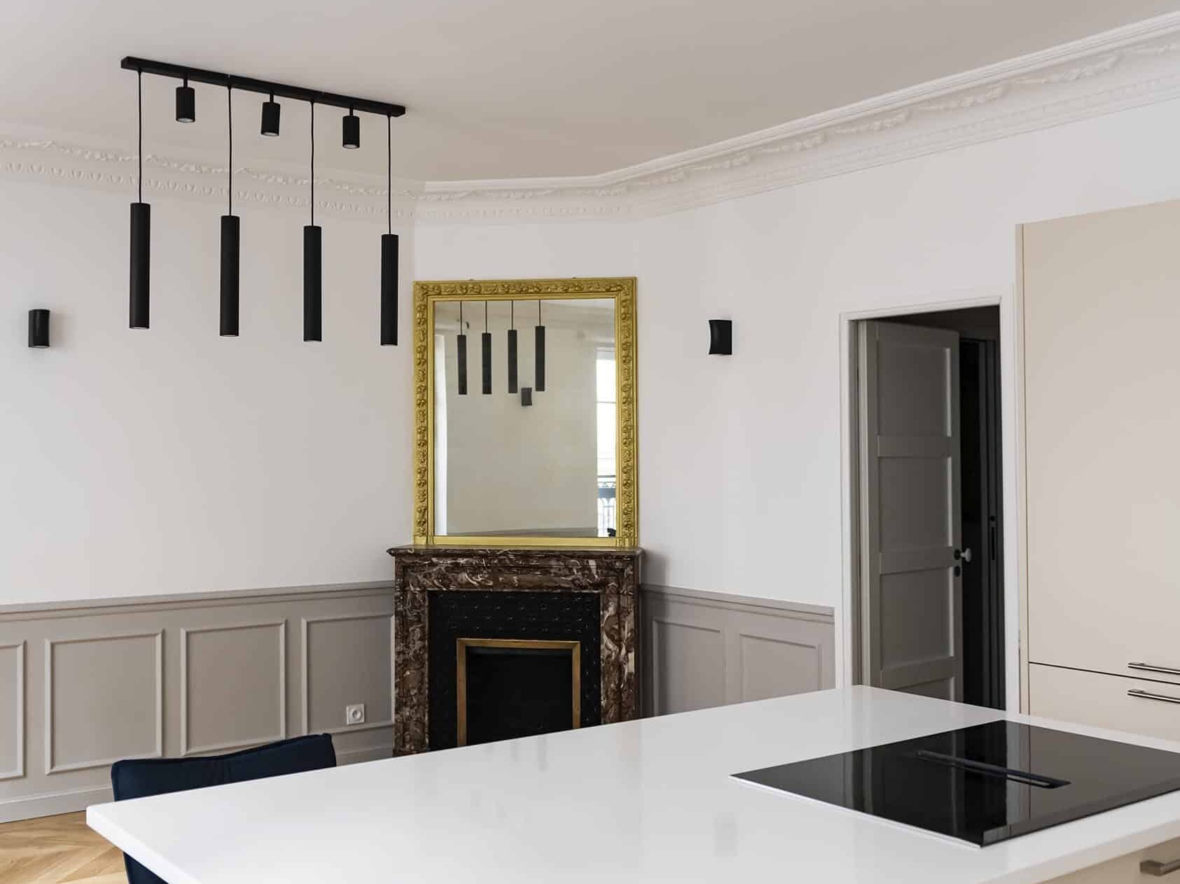 Cuisine moderne de style haussmannien avec îlot blanc, chaises noires et plaque de cuisson électrique. Les caractéristiques comprennent une cheminée avec un miroir au-dessus, des suspensions noires, du parquet et des lambris muraux gris.