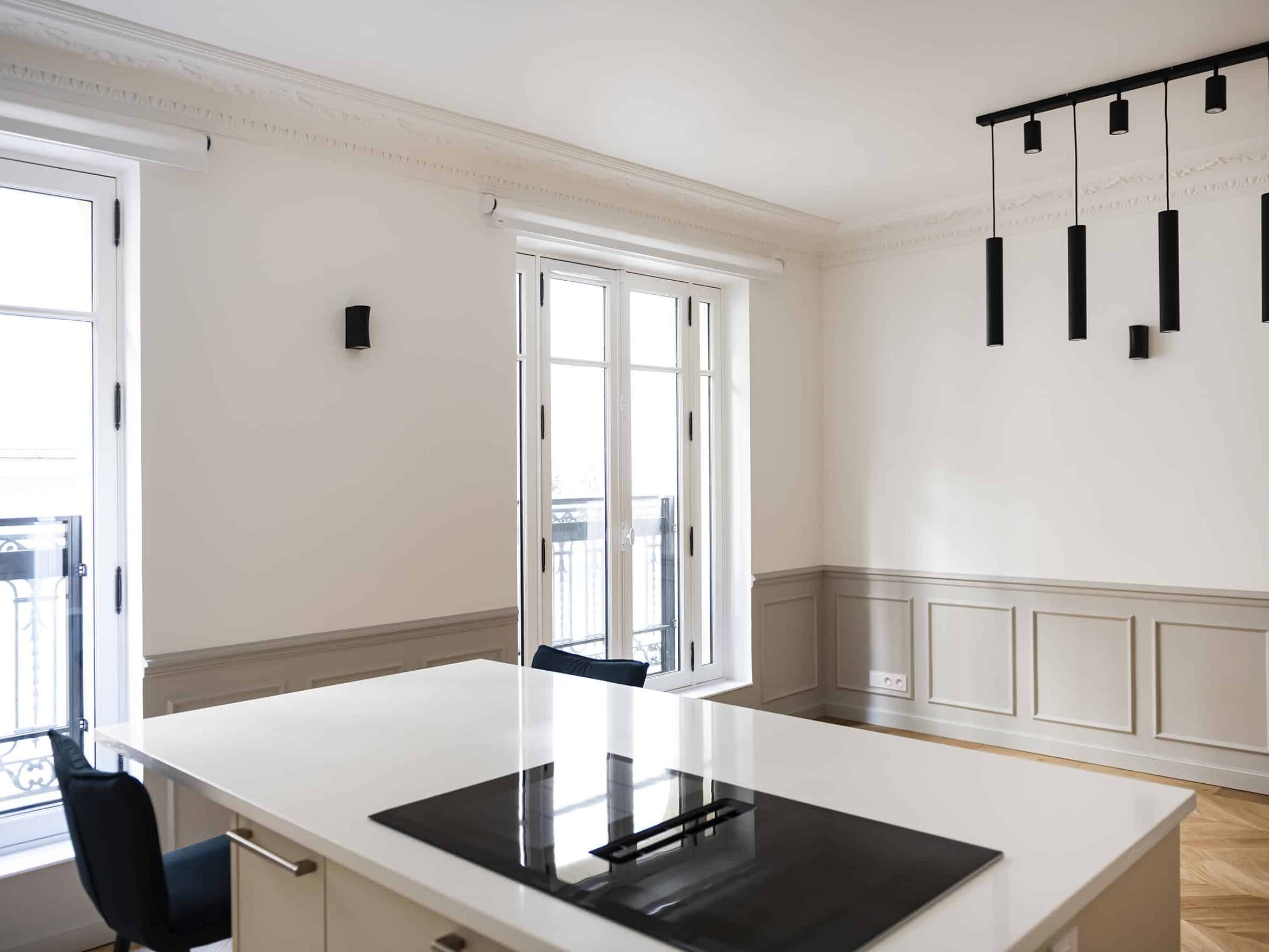 Cette cuisine minimaliste d'inspiration haussmannienne est dotée d'un îlot de cuisine blanc et d'une table de cuisson noire. De grandes fenêtres, un éclairage suspendu noir et d'élégantes moulures murales créent un espace lumineux et moderne.