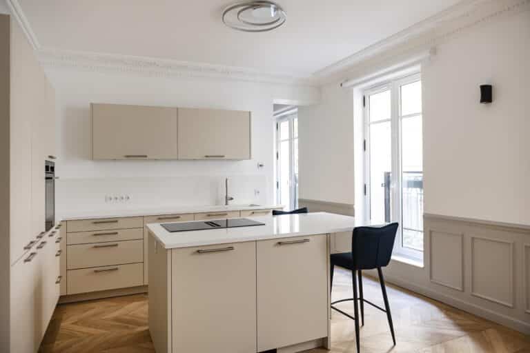 Cette cuisine moderne, rappelant le style haussmannien, est dotée d'armoires beiges, d'un îlot de cuisson à induction avec un tabouret de bar noir et de grandes fenêtres. Les murs blancs et le parquet à chevrons mettent magnifiquement en valeur cet espace lumineux.