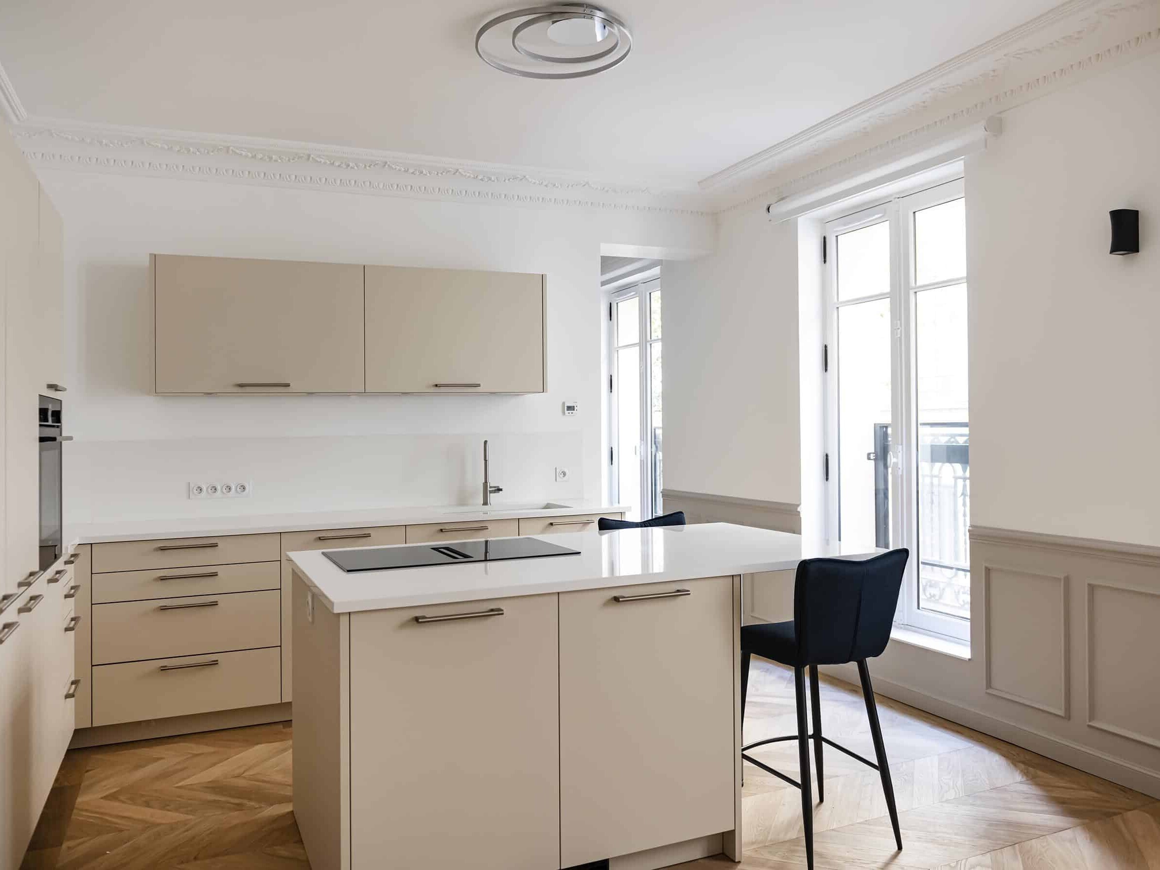 Cette cuisine moderne, rappelant le style haussmannien, est dotée d'armoires beiges, d'un îlot de cuisson à induction avec un tabouret de bar noir et de grandes fenêtres. Les murs blancs et le parquet à chevrons mettent magnifiquement en valeur cet espace lumineux.