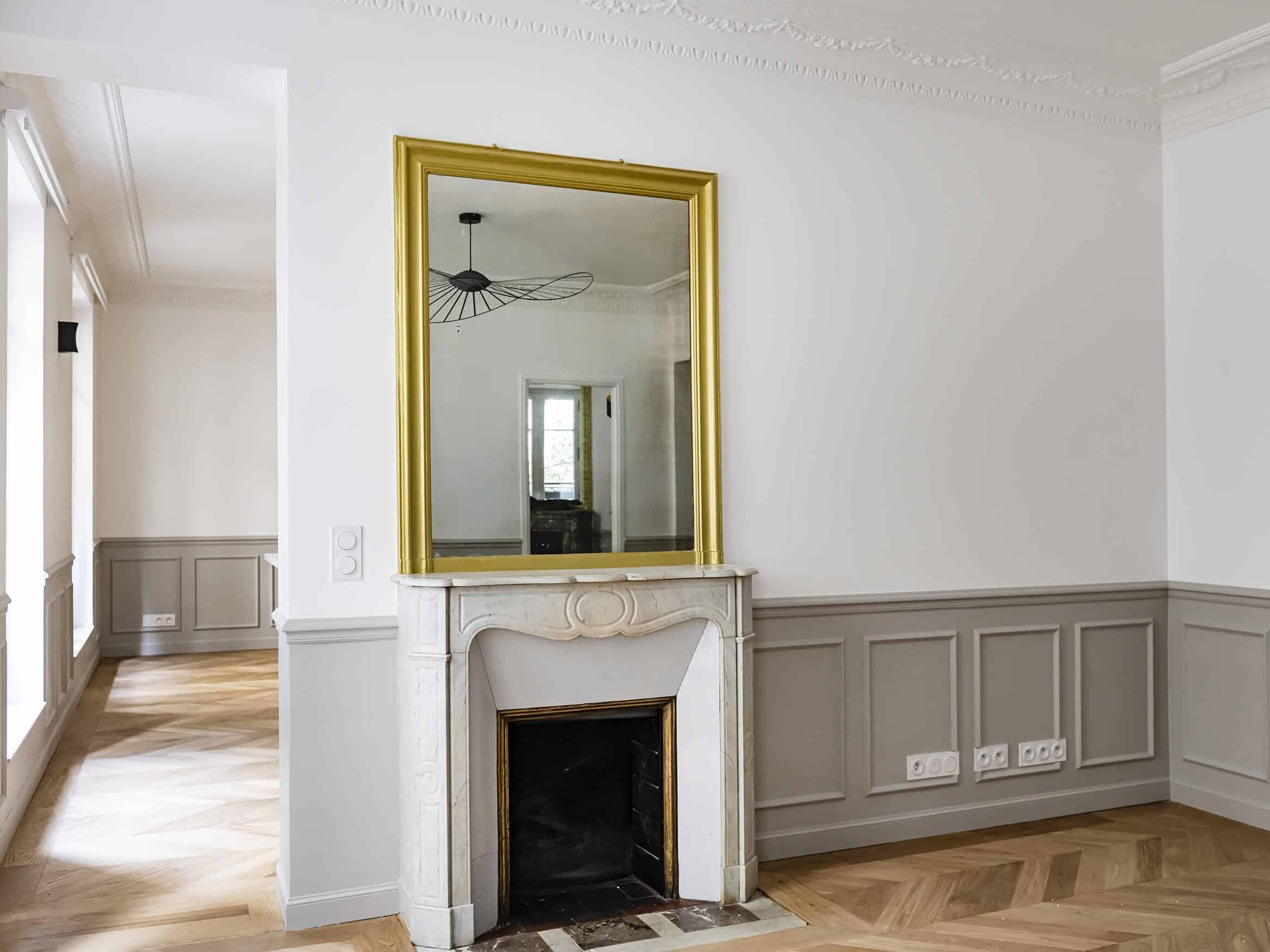 Le charme haussmannien abonde dans cette pièce, avec une cheminée décorative surmontée d'un miroir à cadre doré, des murs lambrissés et un parquet en chevrons. Des moulures au plafond et un luminaire moderne complètent la scène, le tout baigné de lumière naturelle provenant des fenêtres.