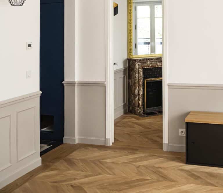 Vue intérieure montrant un couloir avec un parquet en bois à chevrons, des murs blancs et un aperçu d'une pièce avec une cheminée et une fenêtre.